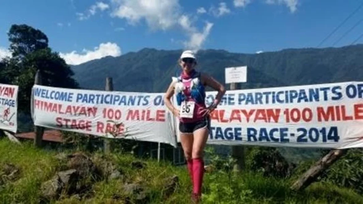 Een vrouw staat naast een spandoek met de tekst "WELKOM DEELNEMERS AAN DE HIMALAYA 100 MILE STAGE RACE - 2014."